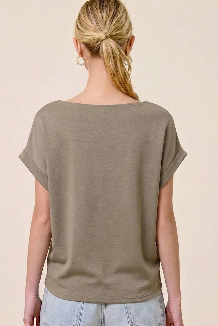 A woman shown from the back wearing a loose, short-sleeve olive-taupe top and light-wash denim shorts against a plain beige studio background.