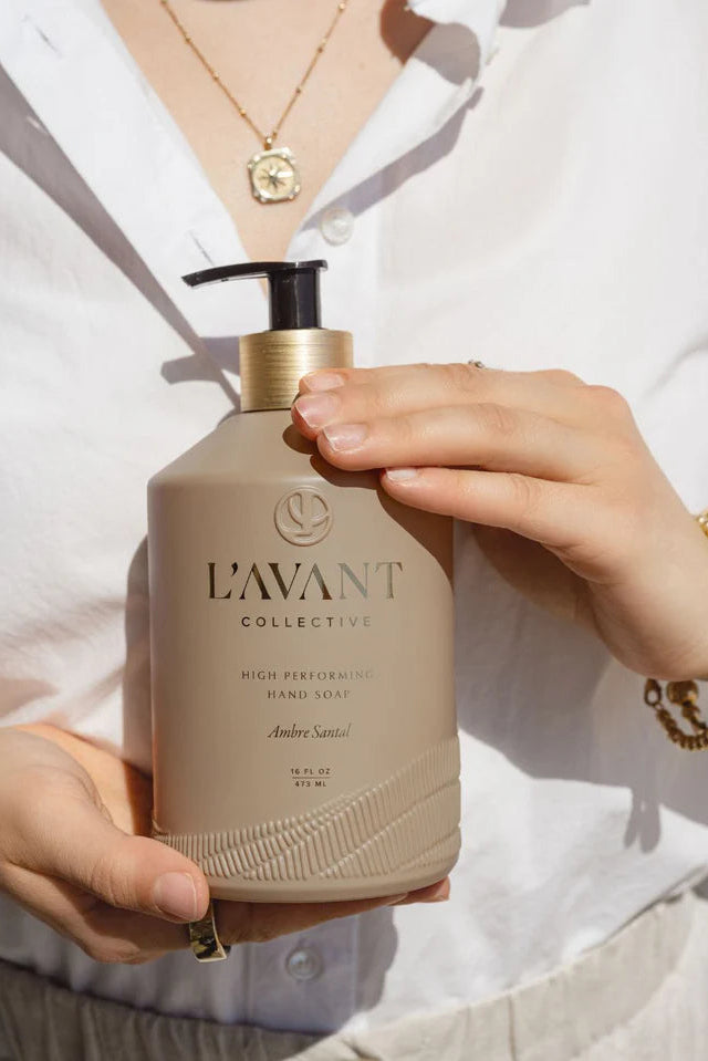 Person holding a beige bottle labeled 'L'AVANT Collective' with a white shirt background