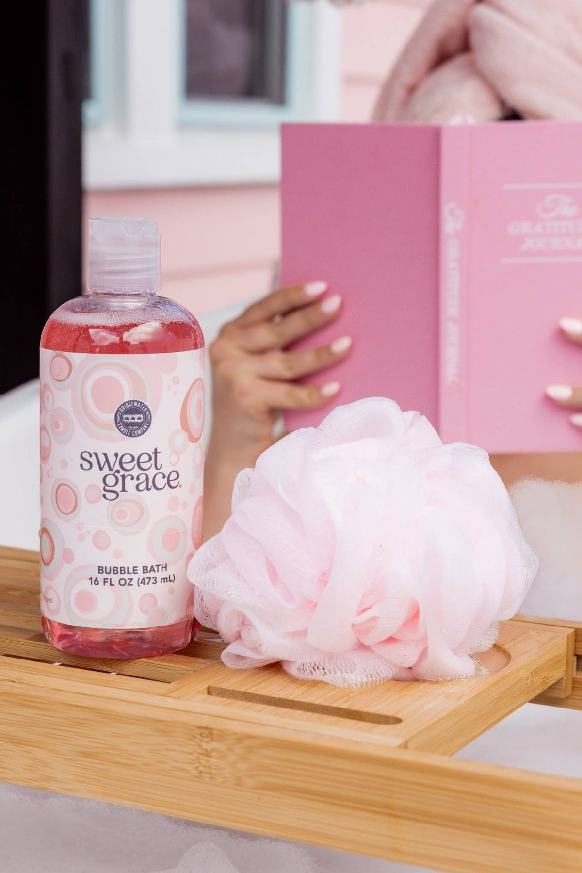 A bottle of Sweet Grace bubble bath, a pink loofah, and a wooden bath caddy in a bubble bath scene with a person holding a pink book in the background.