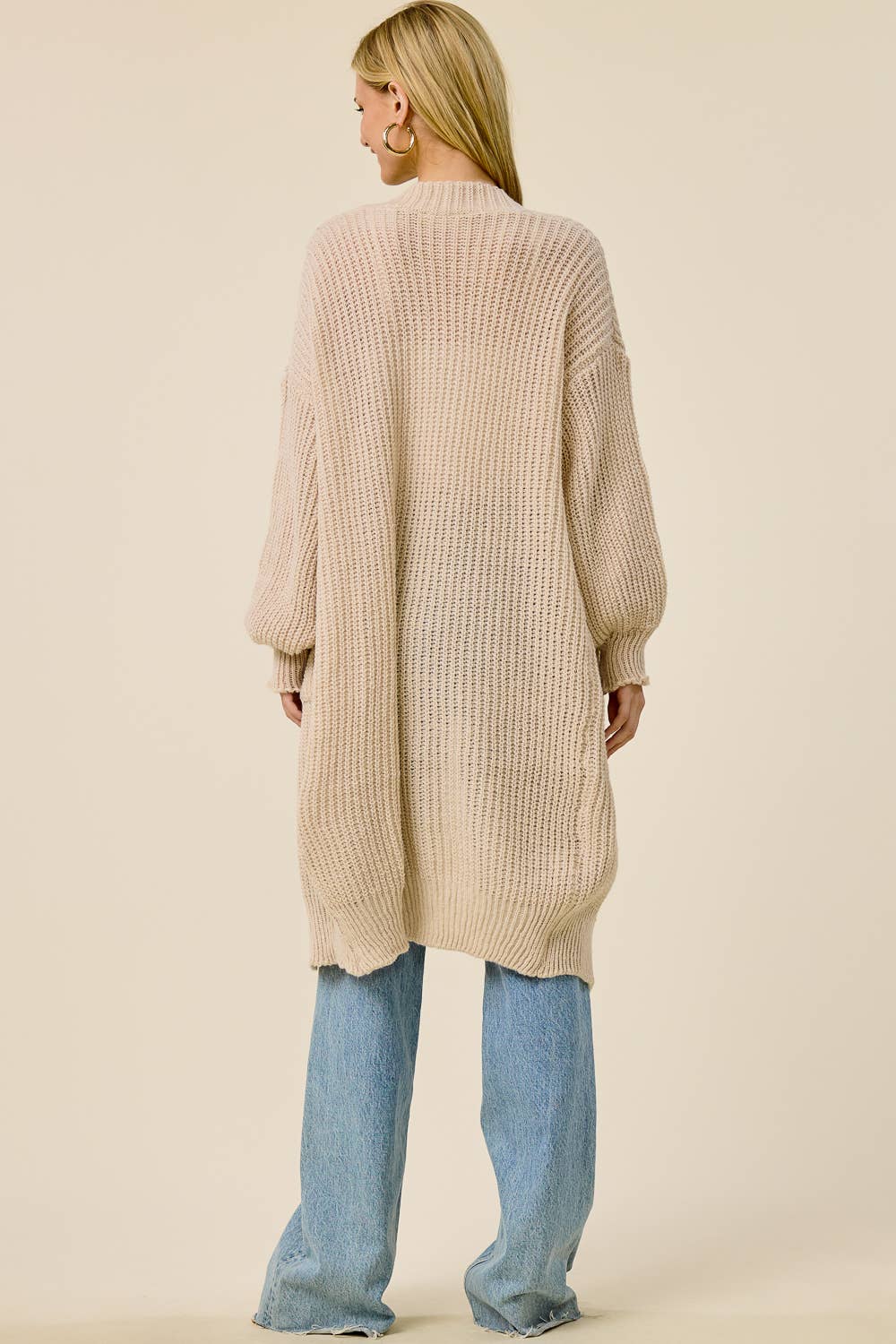 A woman shown from the back wearing a long, oversized ribbed knit cardigan in a light beige color, paired with wide-leg light blue jeans, against a solid cream background.