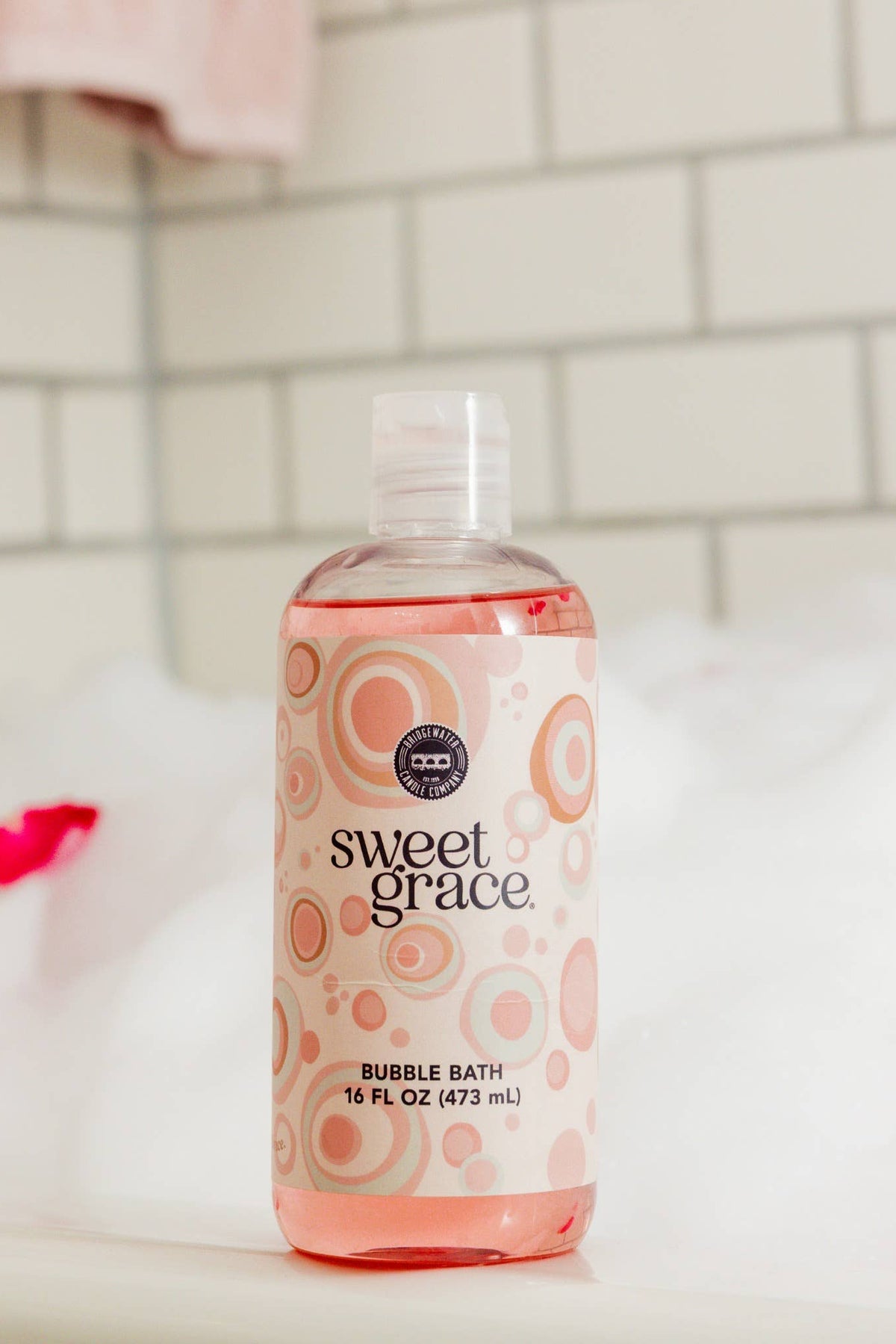 A bottle of Sweet Grace Bubble Bath with pink liquid and a pink label, set on a white tub rim with white bubbles and scattered red rose petals in a tiled bathroom setting.
