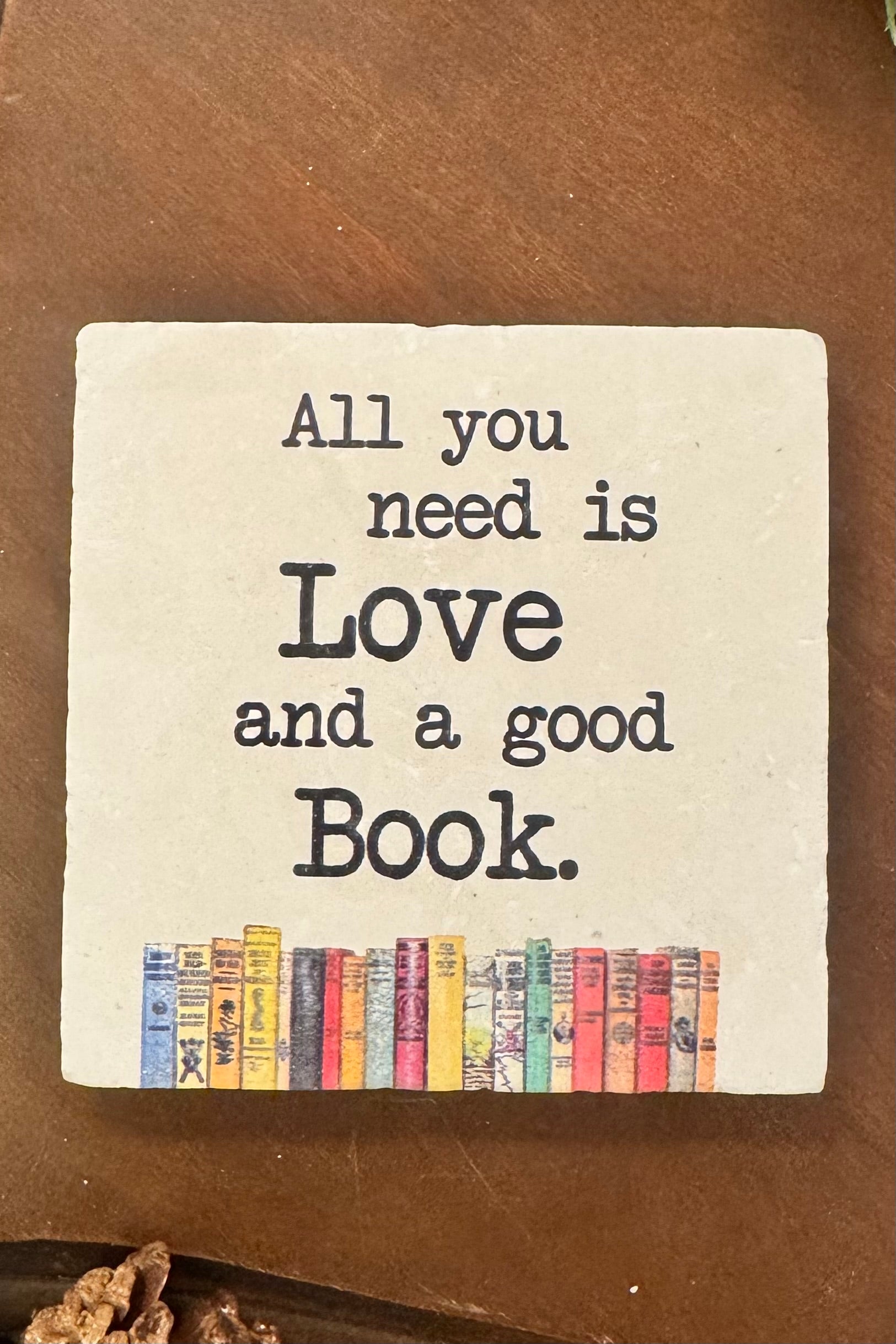 Square coaster with 'All you need is Love and a good Book' text and colorful book illustrations on a brown surface.