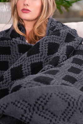A woman wrapped in a charcoal and black fluffy knit throw blanket with a raised geometric diamond pattern, seated on a cream sofa.