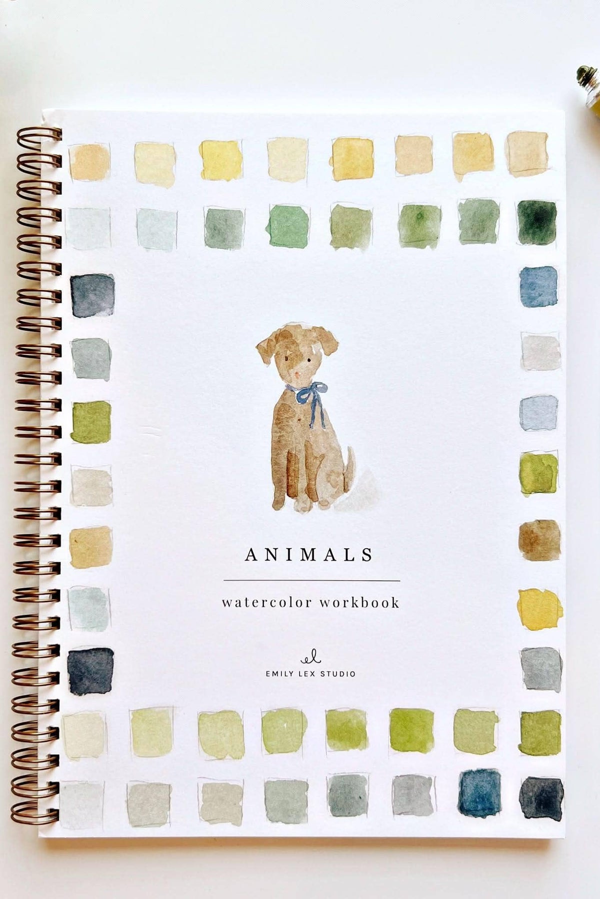 A spiral-bound watercolor workbook titled 'Animals' by Emily Lee Studio, featuring a watercolor illustration of a dog in the center and a border of watercolor color swatches around the edges. The notebook is displayed on a white surface with a paintbrush, a tube of paint, and a green plant leaf.