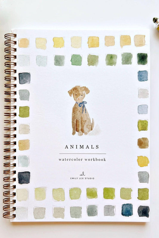 A spiral-bound watercolor workbook titled 'Animals' by Emily Lee Studio, featuring a watercolor illustration of a dog in the center and a border of watercolor color swatches around the edges. The notebook is displayed on a white surface with a paintbrush, a tube of paint, and a green plant leaf.