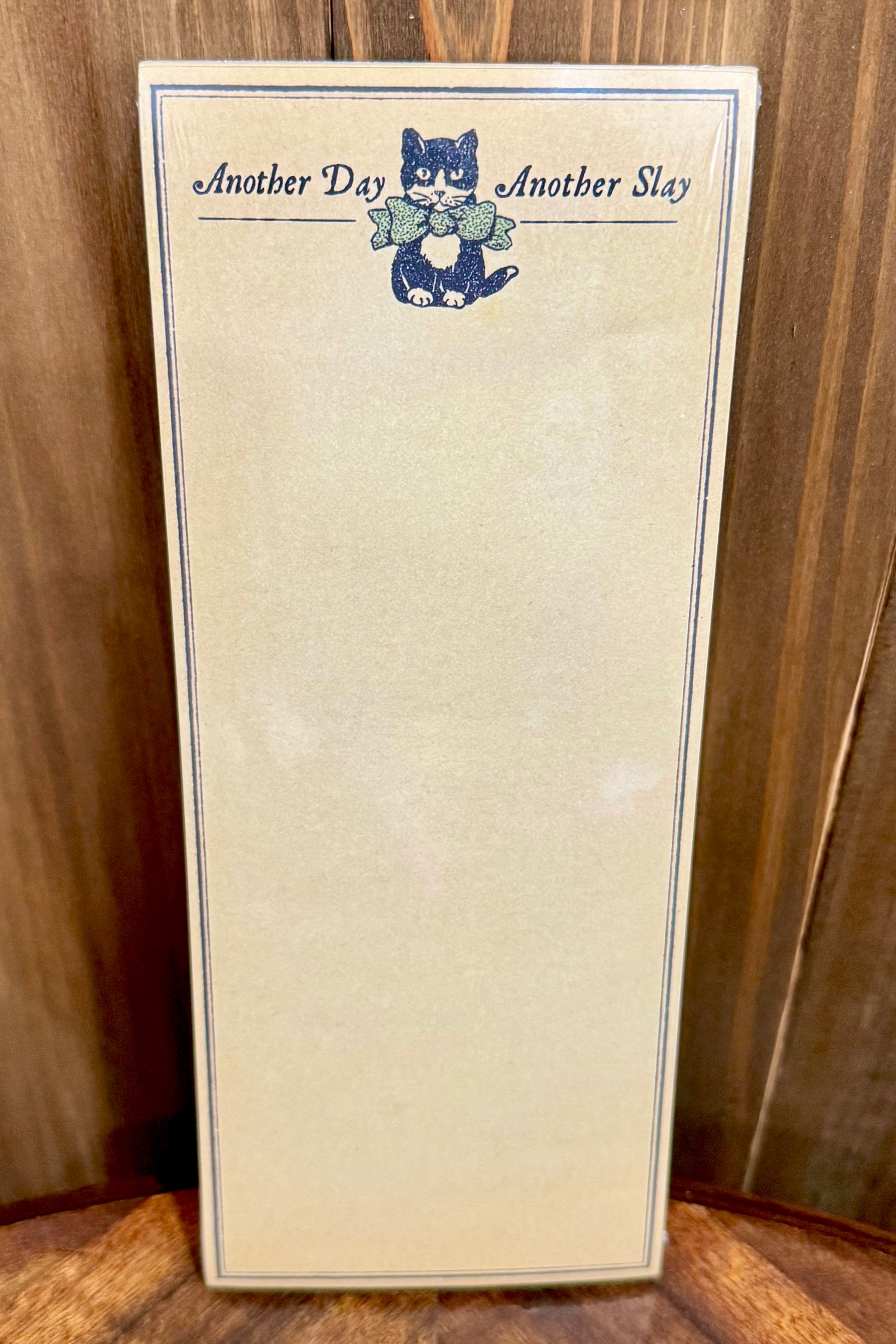 A cream-colored notepad with a blue border, featuring the text 'Another Day Another Slay' and a small illustration of a black and white cat at the top, set against a dark wood background.