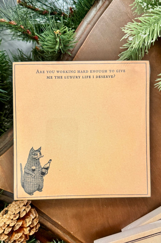 Note card with a cat illustration and humorous text on a brown background, surrounded by Christmas decor.