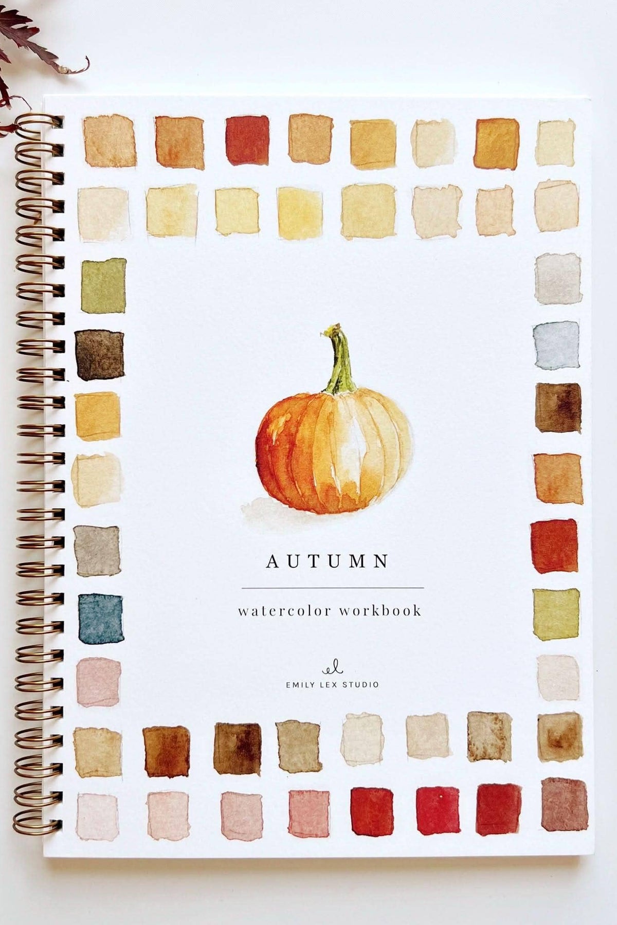 A spiral-bound watercolor workbook titled 'AUTUMN' with a central pumpkin illustration and a border of watercolor color swatches, photographed on a white surface with a paintbrush, tube, and palette visible around it.