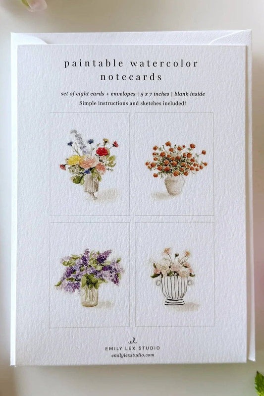 A set of watercolor notecards by Emily Lee Studio displayed on a white surface with a paintbrush, flowers, and greenery. The notecards feature four floral watercolor designs on a white cardstock.