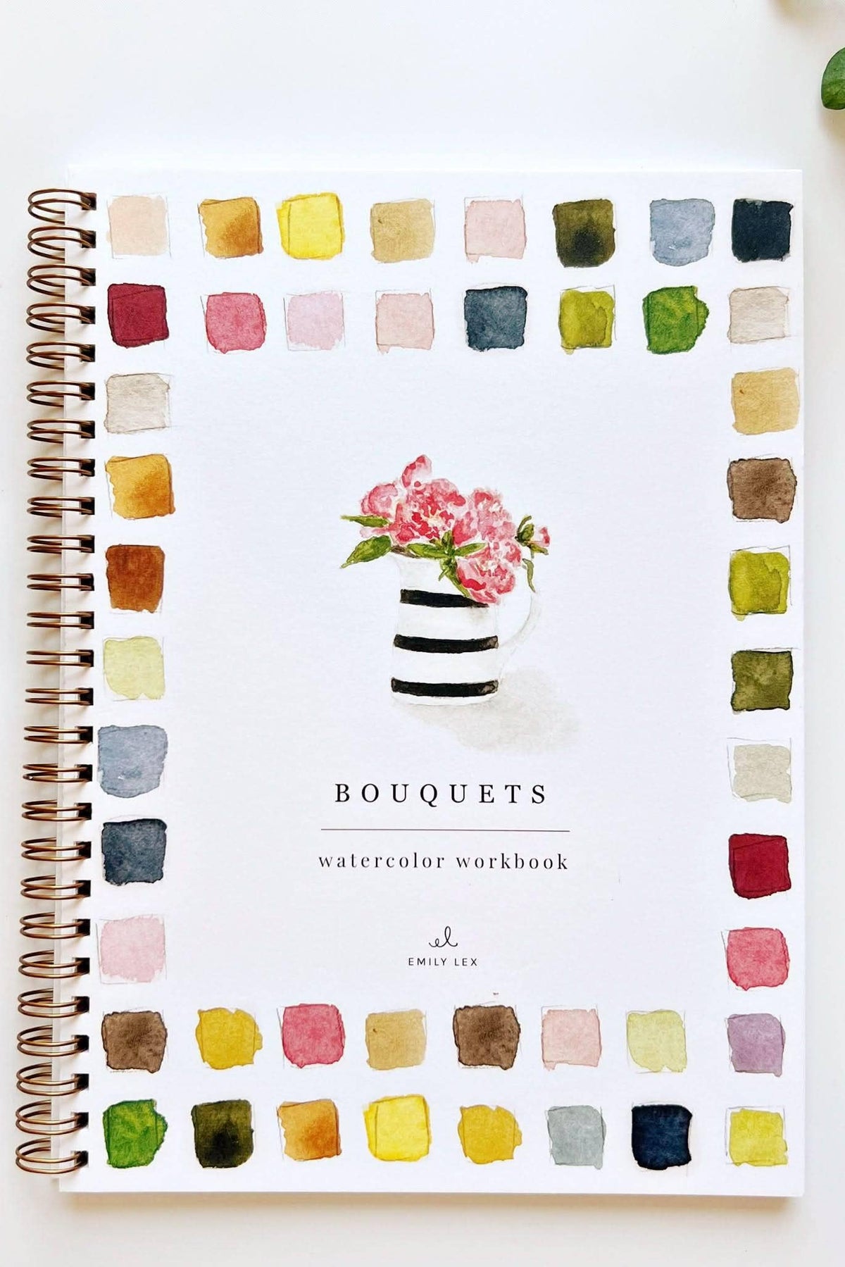 A spiral-bound watercolor workbook titled 'BOUQUETS' with a watercolor flower arrangement on the cover, surrounded by a border of painted color swatches. The workbook is photographed on a white surface with art supplies nearby, including a paint tube, paintbrush, and small plant.