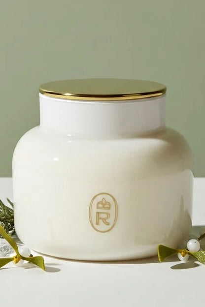 A white ceramic jar with a gold lid and a gold monogram logo on the front, styled with green leaves and white pearls on a white surface against a sage green background.