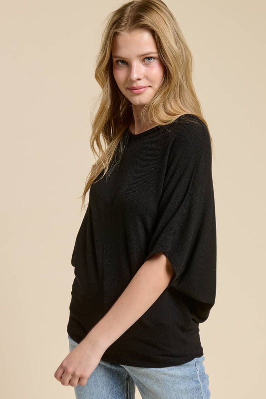 A woman modeling a black short-sleeve knit top with a relaxed, draped fit, paired with light blue jeans, against a solid beige studio background.