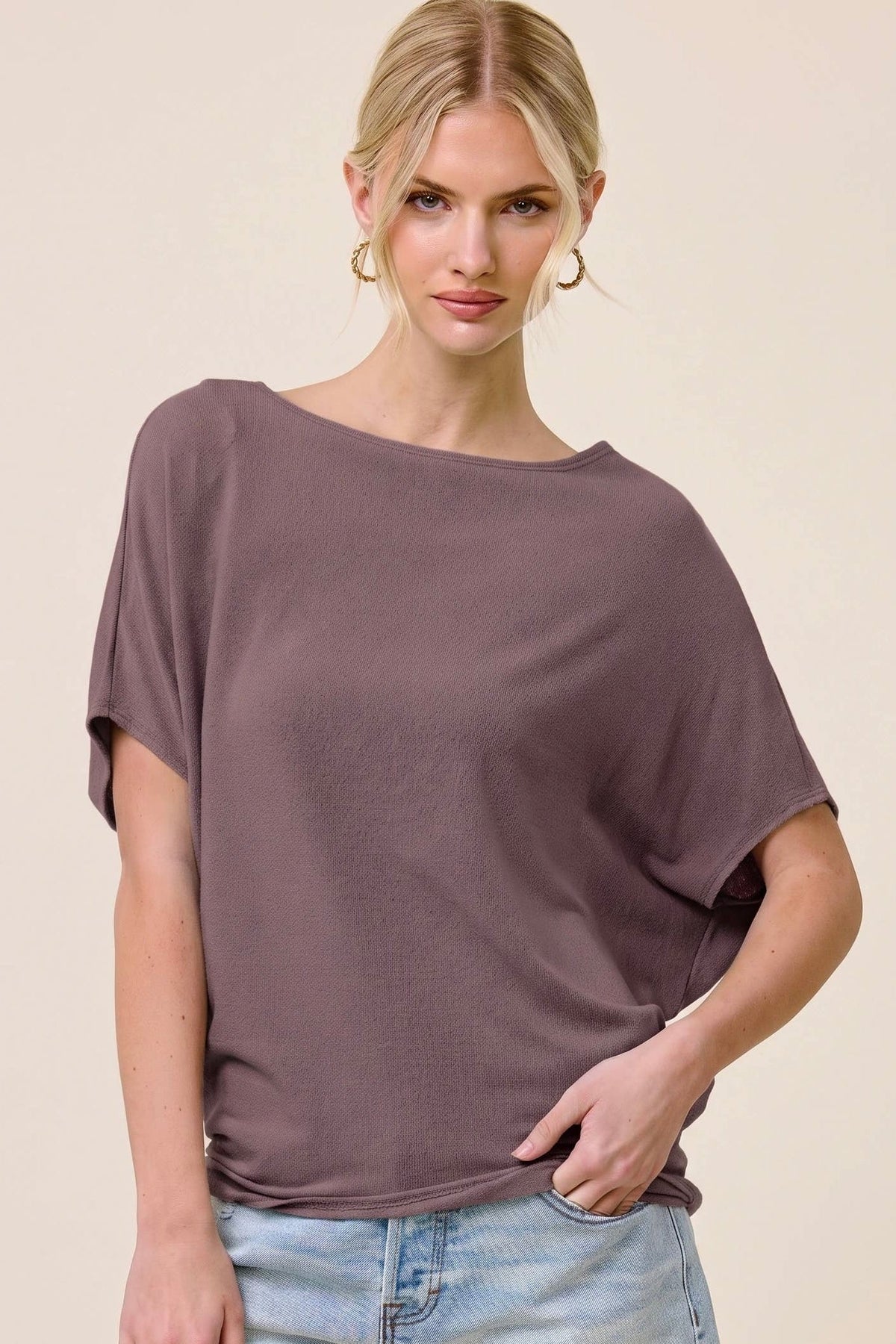 A woman modeling a loose, short-sleeve boat-neck top in a muted mauve-purple color, paired with light-wash denim shorts, against a solid beige studio background.