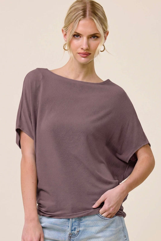 A woman modeling a loose, short-sleeve boat-neck top in a muted mauve-purple color, paired with light-wash denim shorts, against a solid beige studio background.