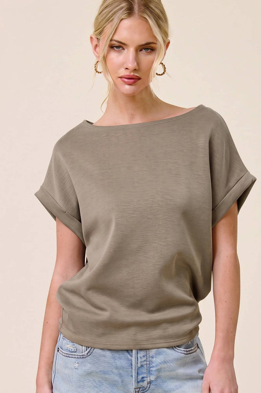 A woman modeling a loose, short-sleeve knit top in taupe/olive-gray, paired with light-wash distressed denim shorts, against a plain beige studio background.