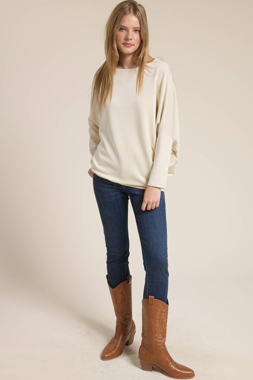 Woman wearing a cream sweater, blue jeans, and brown boots on a plain background