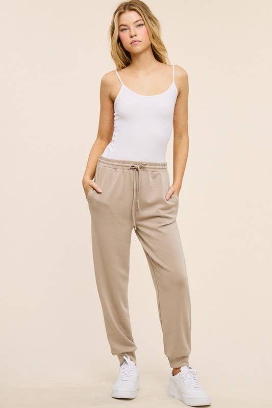 Woman wearing taupe sweatpants and a white tank top on a beige background