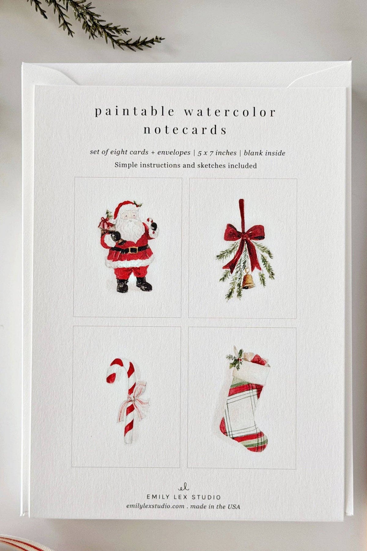 A flat-lay of a notepad card set titled 'paintable watercolor notecards' by Emily Lee Studio, featuring four sample watercolor illustrations of Santa Claus, mistletoe, a candy cane, and a Christmas stocking. The card is styled on a white surface with a paintbrush, evergreen branches, and striped fabric.