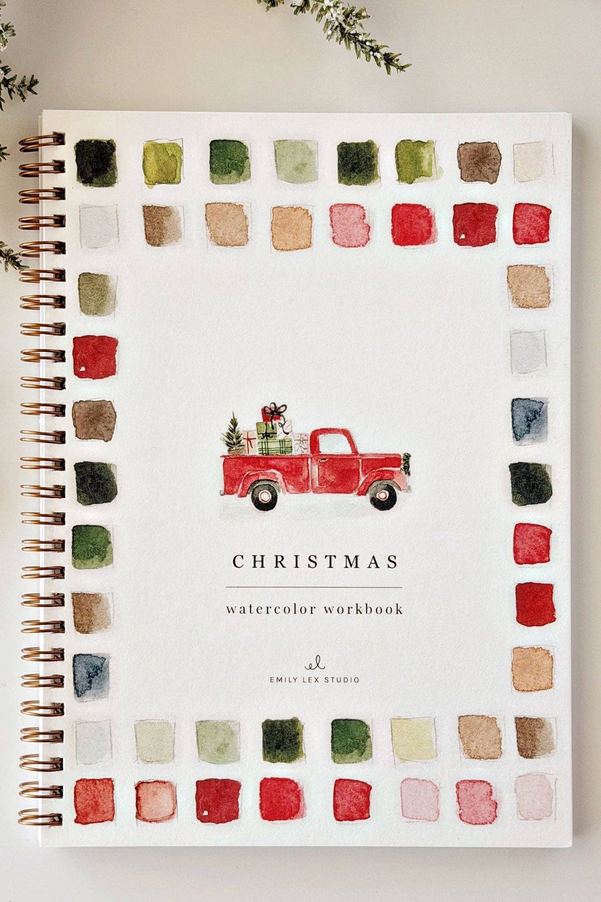 A spiral-bound watercolor workbook titled 'CHRISTMAS watercolor workbook' by Emily Lee Studio, styled in a flat lay with a red truck illustration and watercolor swatches around the border. A paint tube, paintbrush, and greenery are arranged around it on a light background.