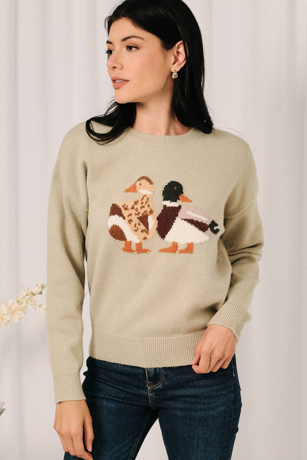 A woman wearing a beige crewneck sweater with a knitted duck design on the front, paired with dark blue jeans, standing against a light curtain background.