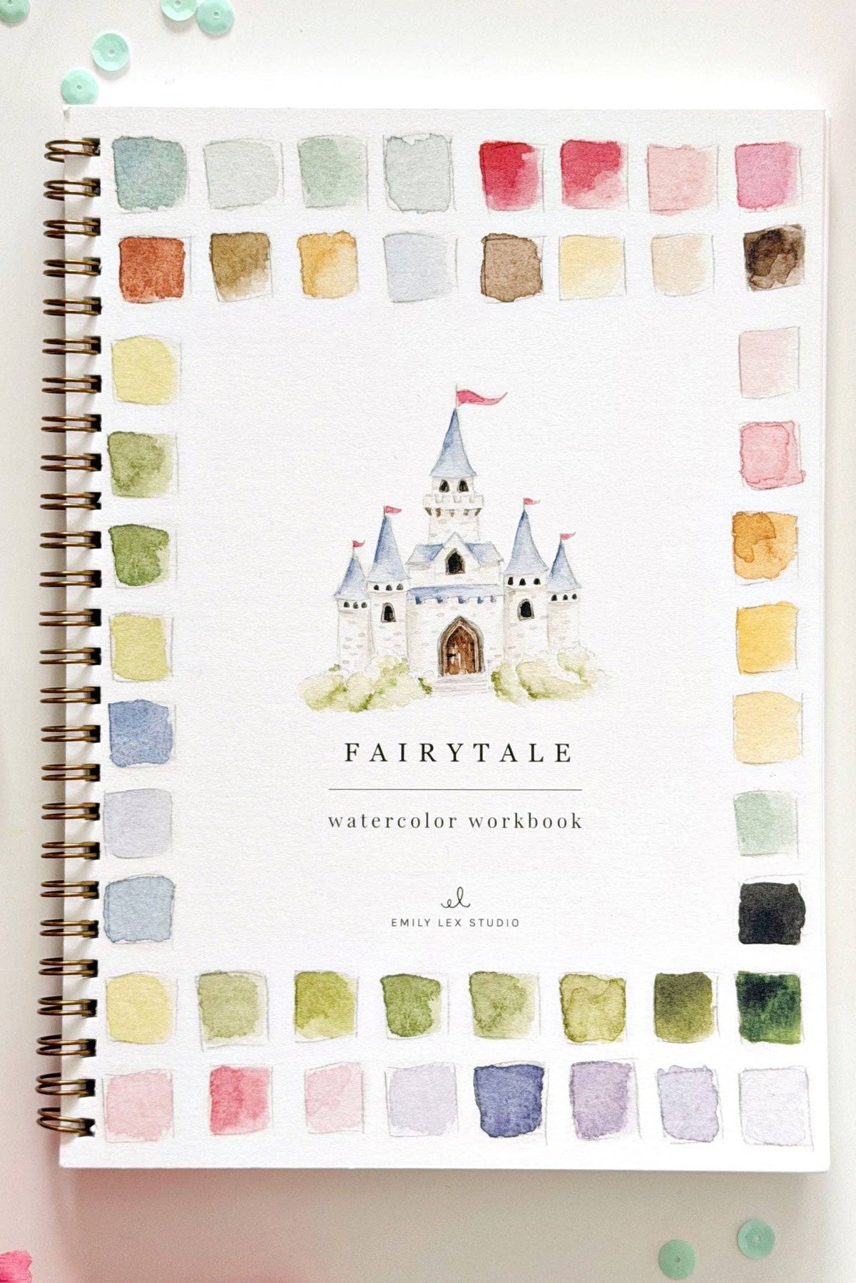 A spiral-bound watercolor workbook titled 'FAIRYTALE' with a castle illustration on the cover, surrounded by watercolor swatches in a border layout. The book is photographed on a white surface with decorative flowers, beads, a paintbrush, and ribbon.