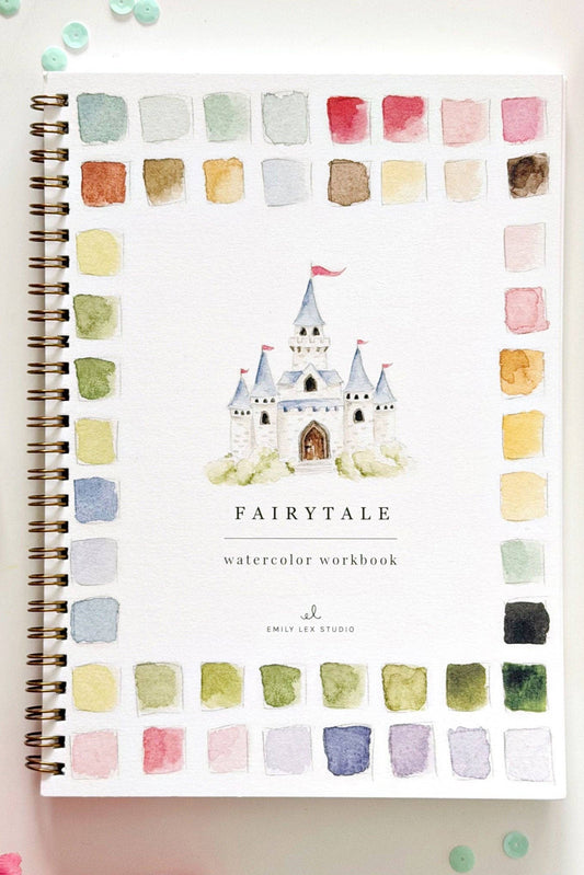 A spiral-bound watercolor workbook titled 'FAIRYTALE' with a castle illustration on the cover, surrounded by watercolor swatches in a border layout. The book is photographed on a white surface with decorative flowers, beads, a paintbrush, and ribbon.