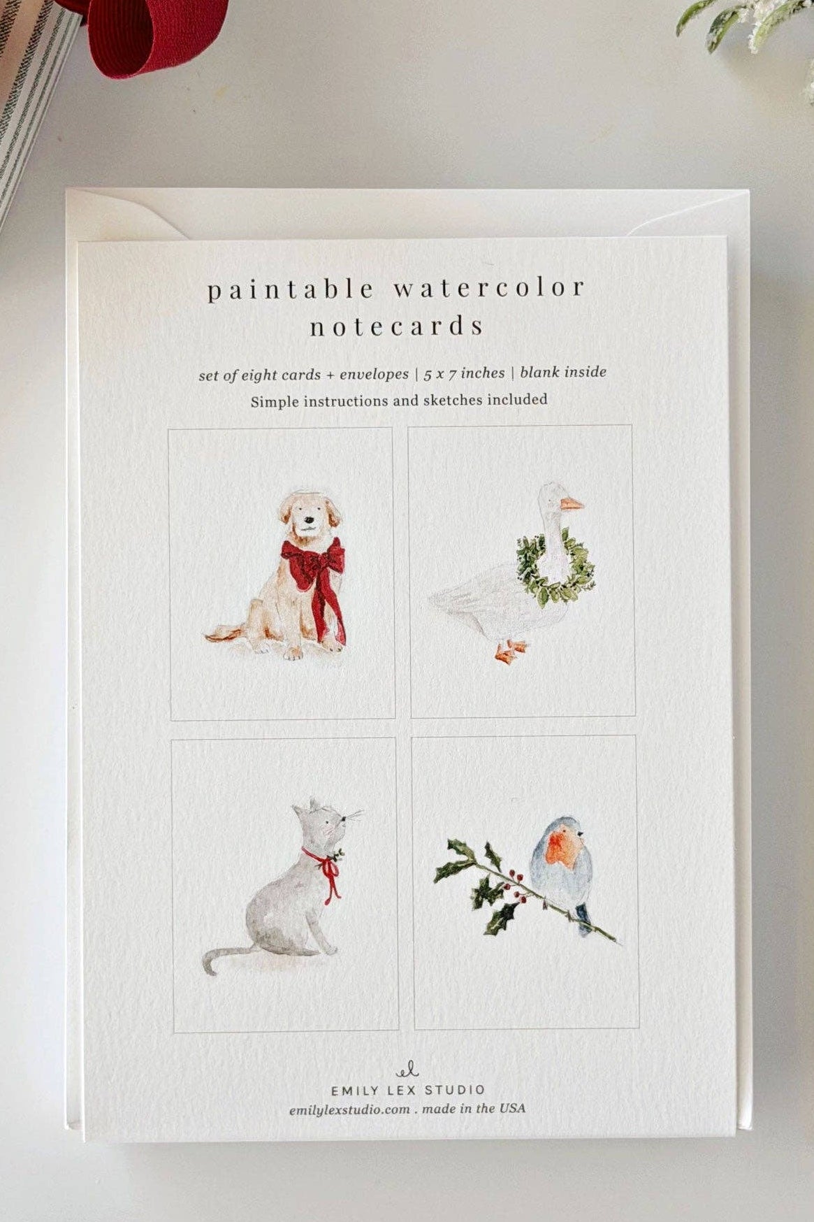 A flat-lay of a set of eight printable watercolor notecards by Emily Levy Studio, displayed on a white surface with a red ribbon, a paintbrush, and a sprig of white flowers.