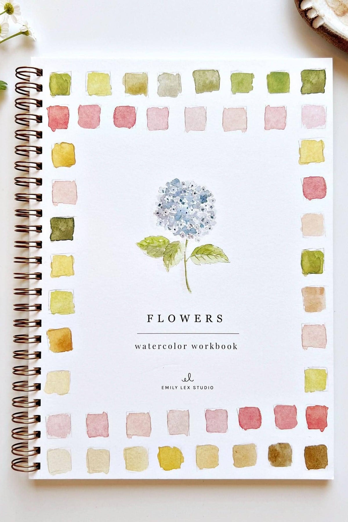A spiral-bound watercolor workbook titled 'FLOWERS' lies on a white surface, surrounded by a watercolor border of pastel flower-like swatches. A paintbrush rests to the right, with small white flowers and a plate of fruit partially visible at the edges.
