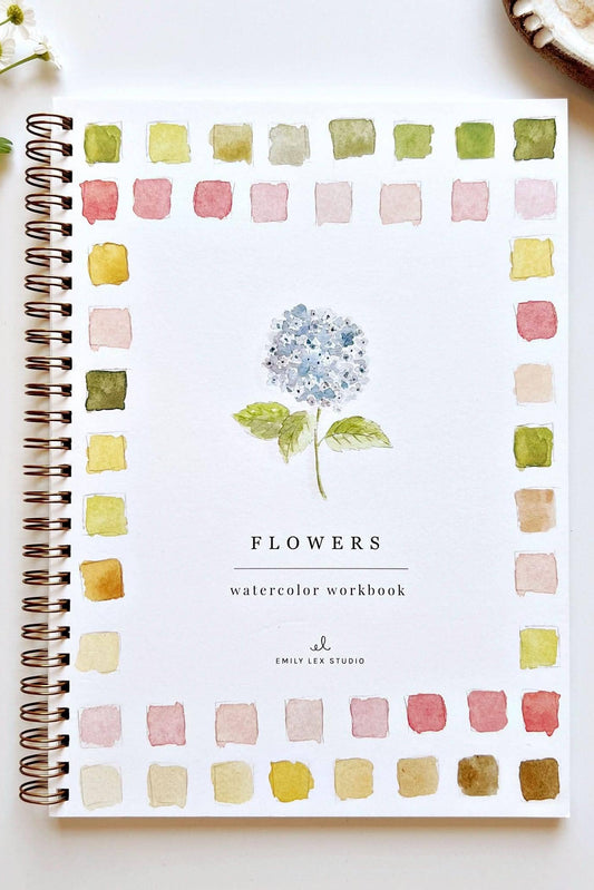A spiral-bound watercolor workbook titled 'FLOWERS' lies on a white surface, surrounded by a watercolor border of pastel flower-like swatches. A paintbrush rests to the right, with small white flowers and a plate of fruit partially visible at the edges.