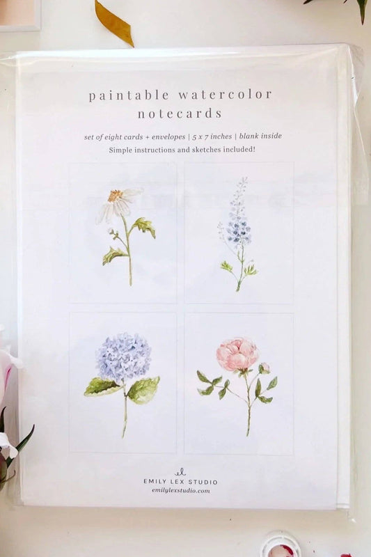 A flat-lay of a watercolor floral notepad set on a white surface with flowers and a paintbrush. The notepad reads 'paintable watercolor notecards' and shows four watercolor flower illustrations.