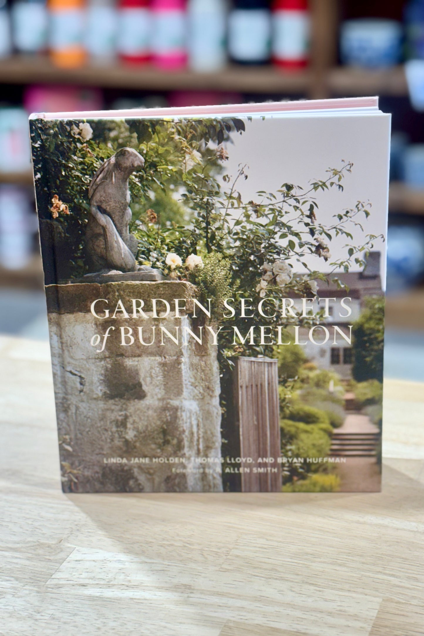 Book titled 'Garden Secrets of Bunny Mellon' on a wooden surface with blurred shelves in the background.