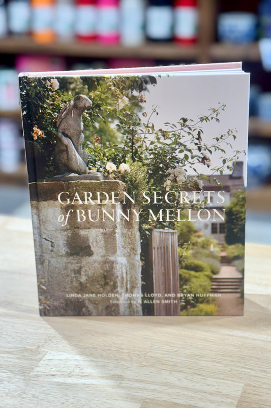Book titled 'Garden Secrets of Bunny Mellon' on a wooden surface with blurred shelves in the background.