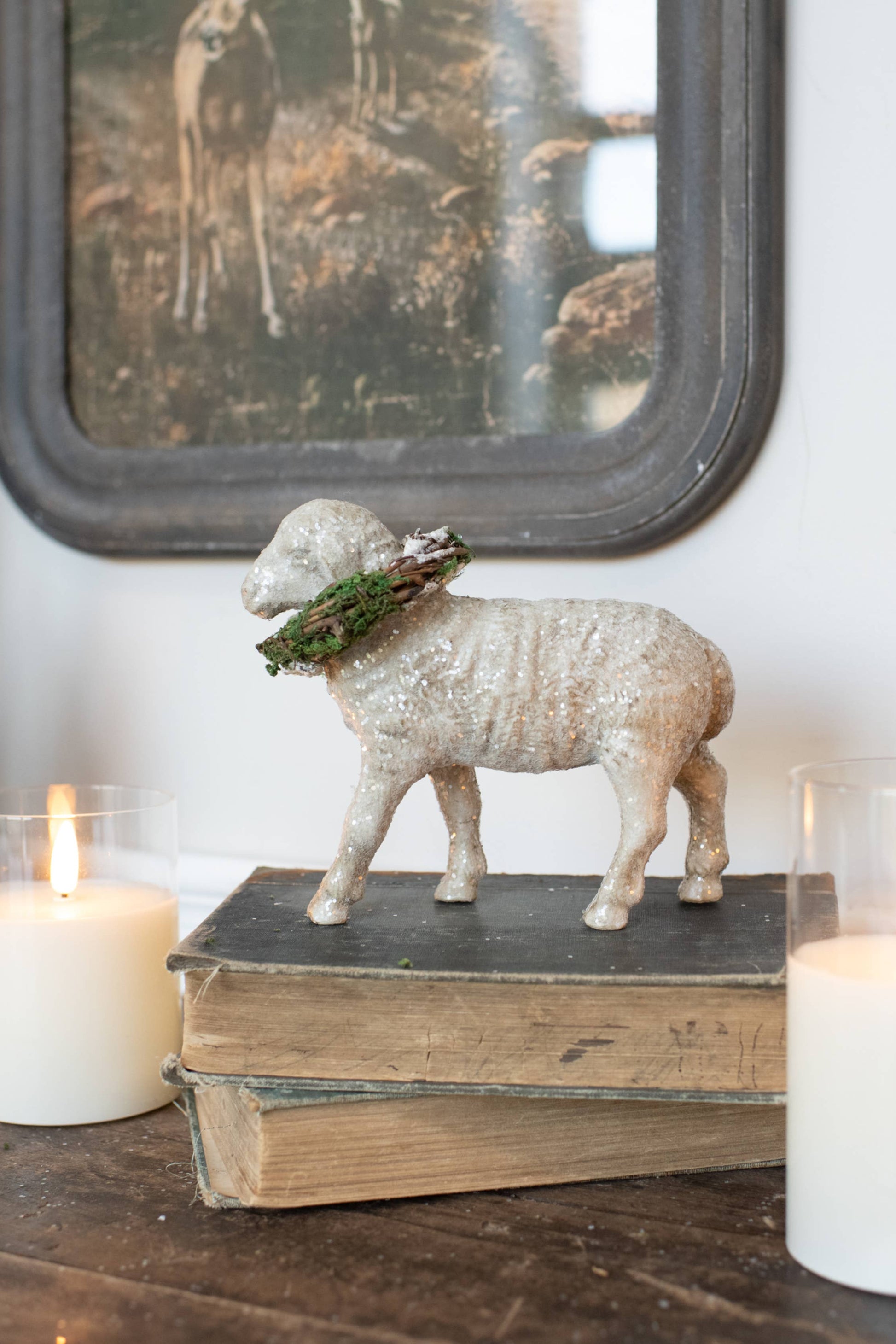 Decorative setup with sheep figurine, candles, and books on a wooden surface.