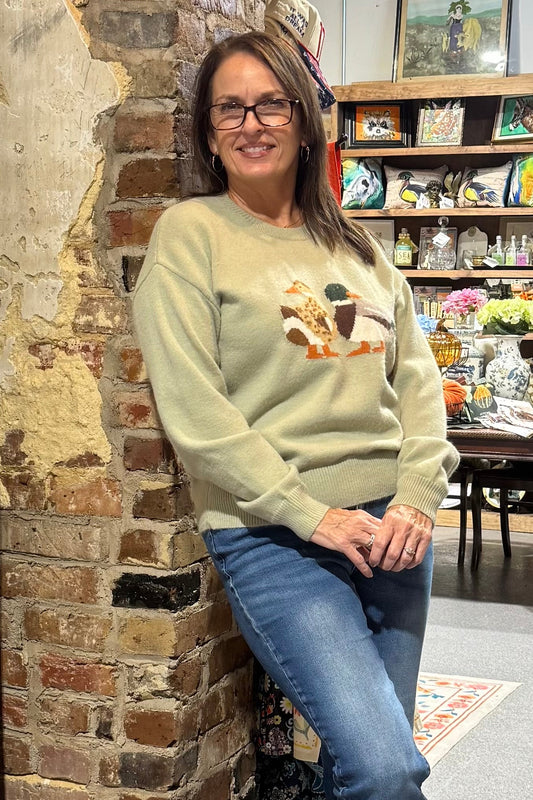 A woman wearing a light sage green crewneck sweater with a colorful mallard duck design on the chest, paired with blue jeans and white sneakers, leaning against a brick pillar in a rustic shop setting.