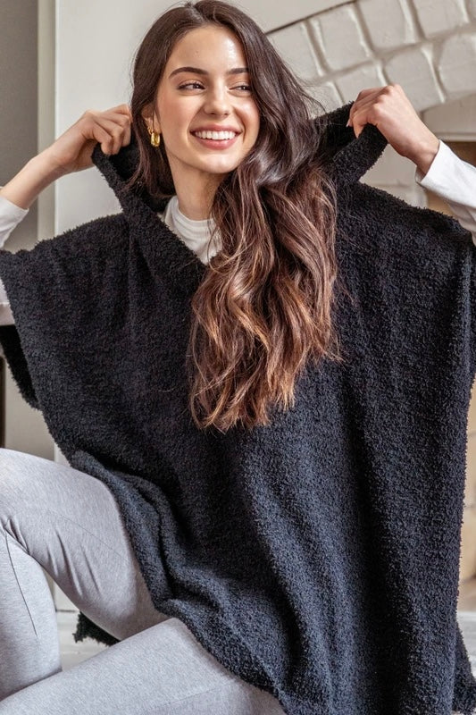A woman wearing a black fuzzy hooded poncho sweater, holding the hood up with both hands. She is sitting indoors with a light-colored brick wall in the background.