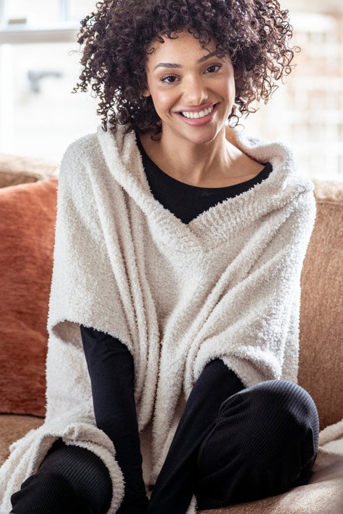 A smiling woman with curly dark hair sits on a couch wearing a cream-colored, plush, textured poncho-style sweater over a black top and black pants.