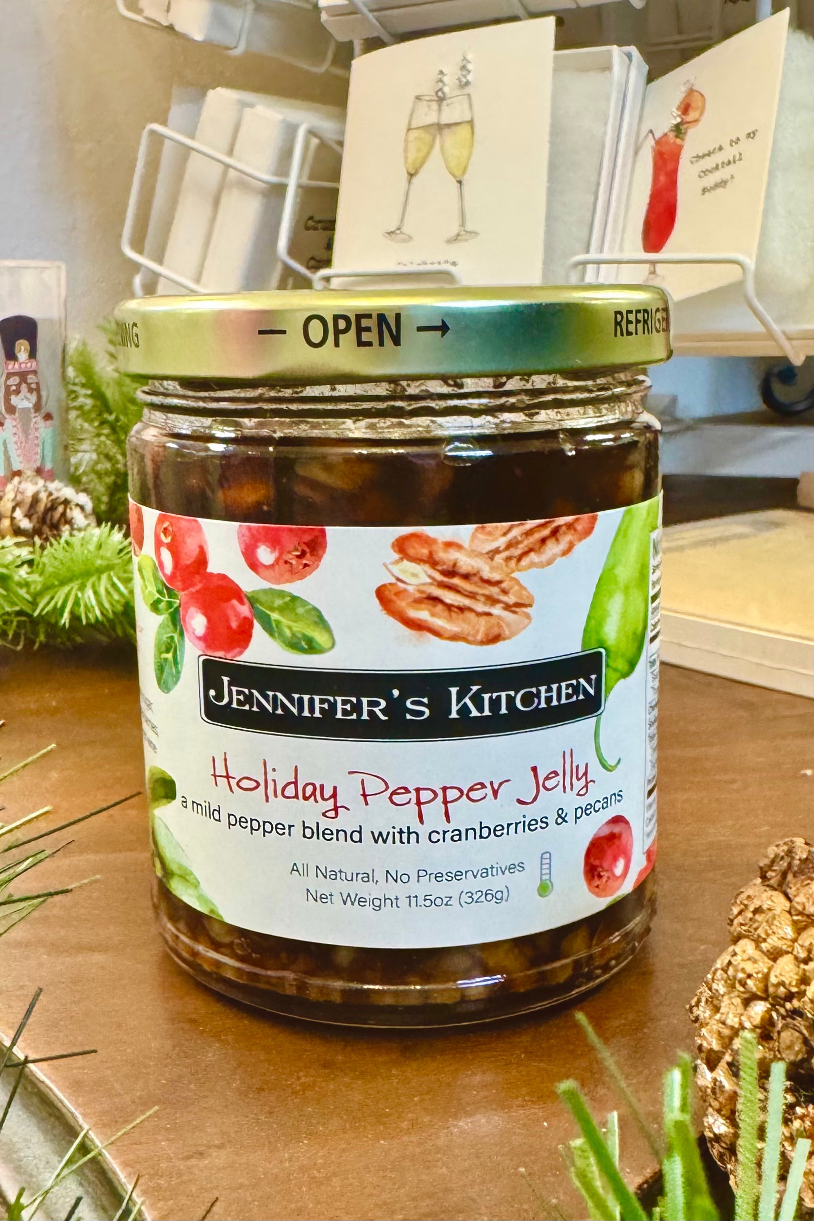 Jar of Jennifer's Kitchen Holiday Pepper Jelly on a wooden surface with decorative items in the background.