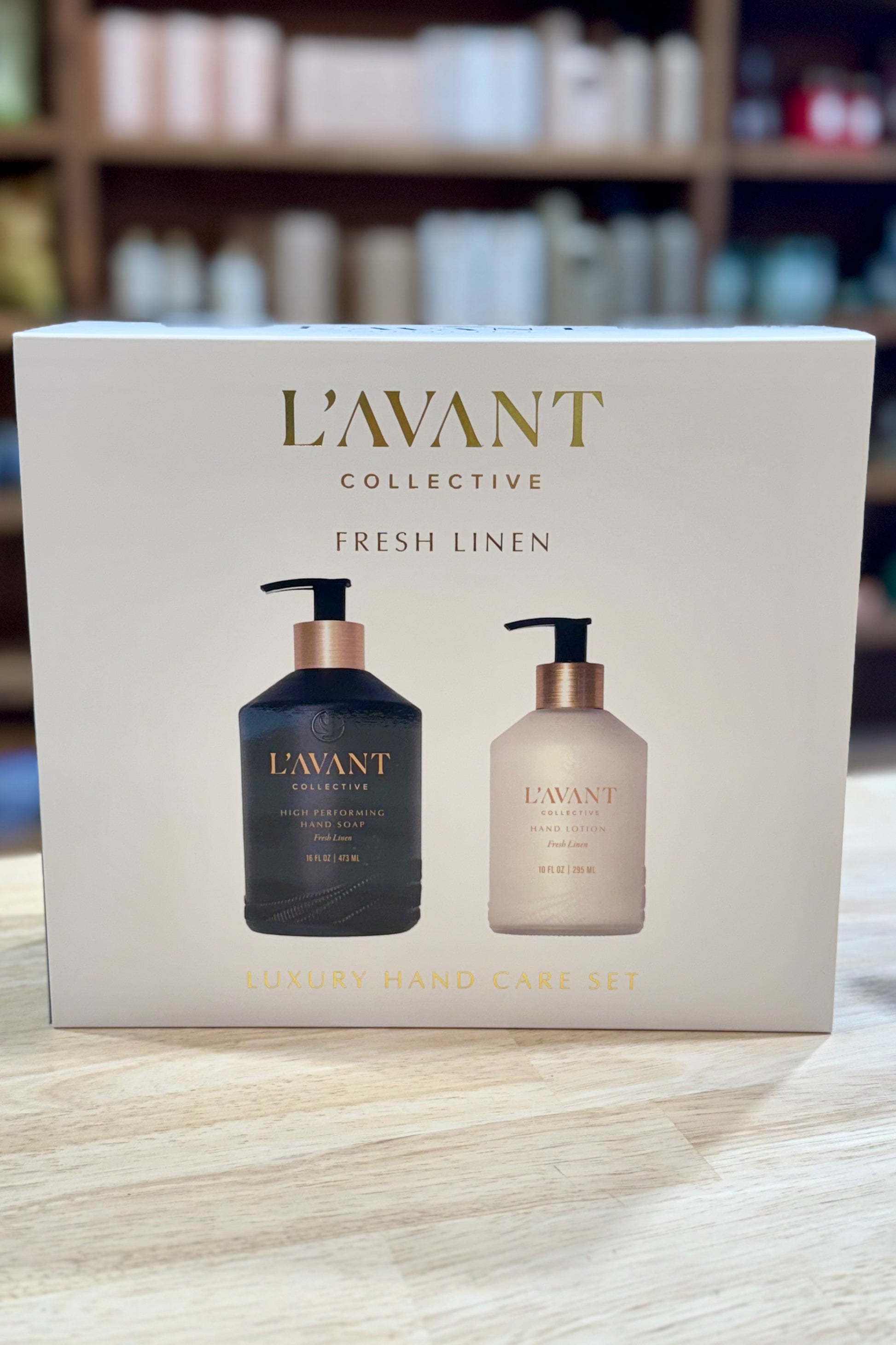 L'Avant Collective hand care set packaging on a wooden surface with blurred shelves in the background.