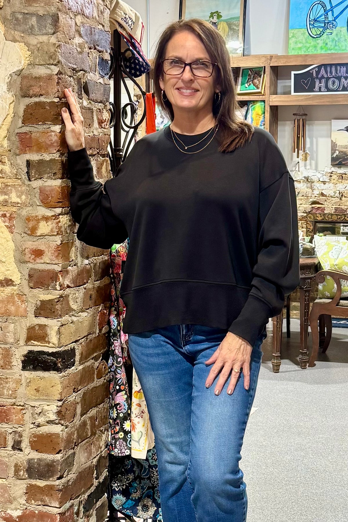 A woman with glasses and brown hair, wearing a black crewneck sweatshirt and blue jeans, posing next to a brick wall in a shop setting.