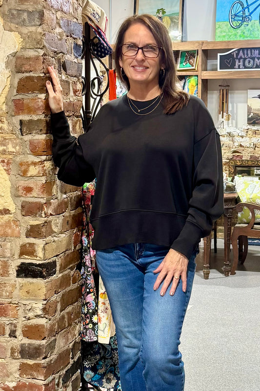A woman with glasses and brown hair, wearing a black crewneck sweatshirt and blue jeans, posing next to a brick wall in a shop setting.