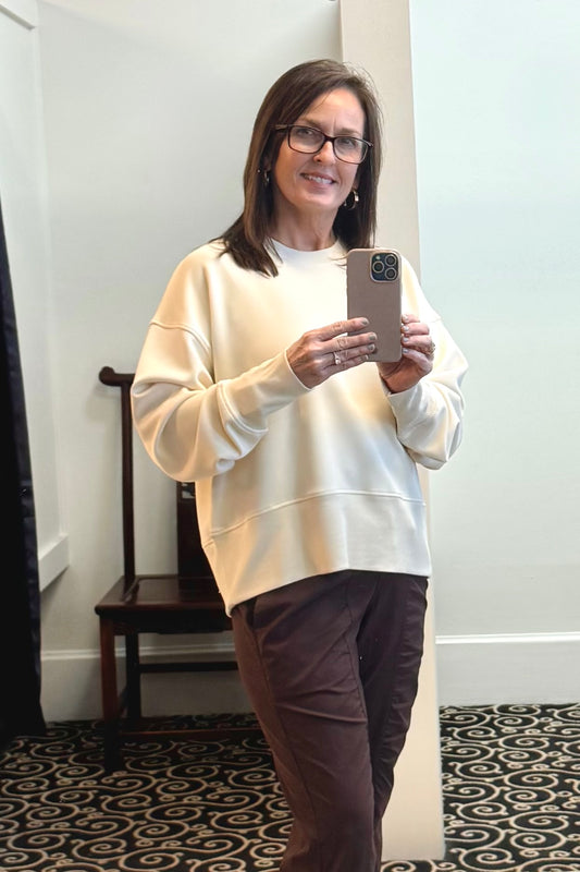 Woman taking a mirror selfie wearing a cream sweatshirt and brown pants in a room with a chair and patterned carpet.