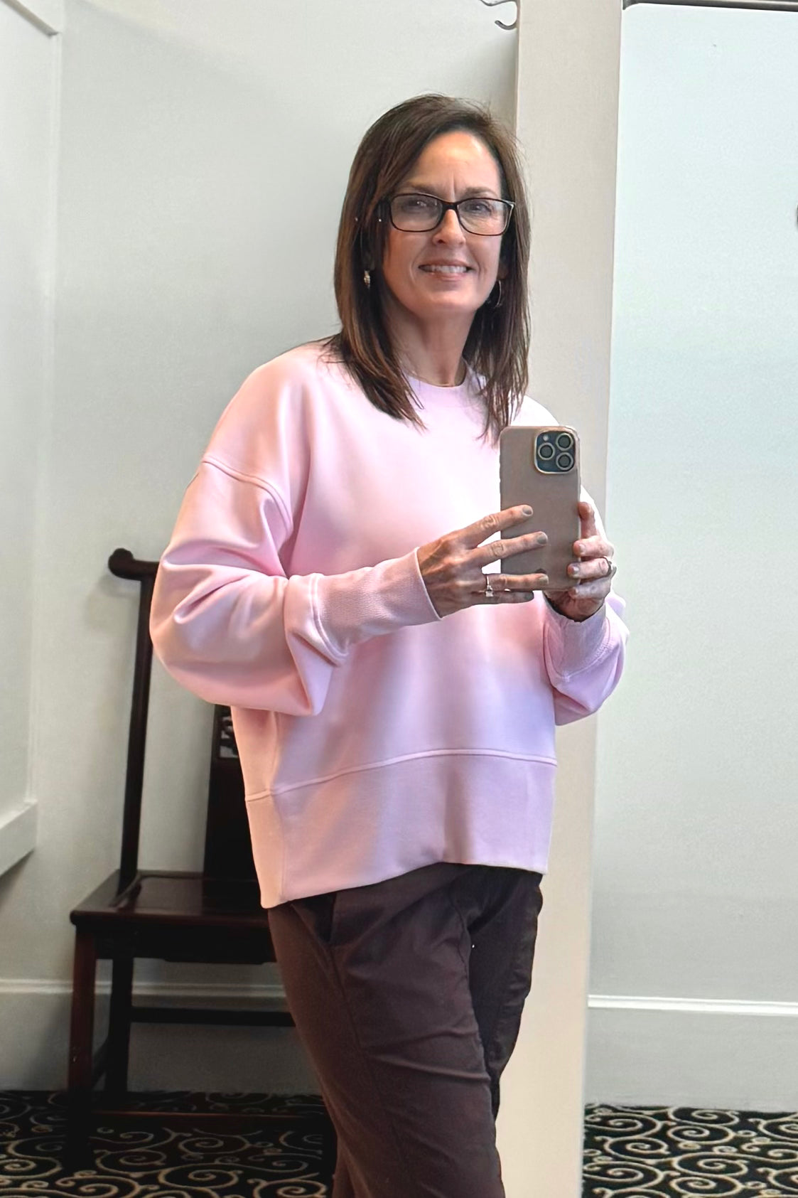 Woman taking a mirror selfie wearing a pink sweatshirt and glasses indoors.