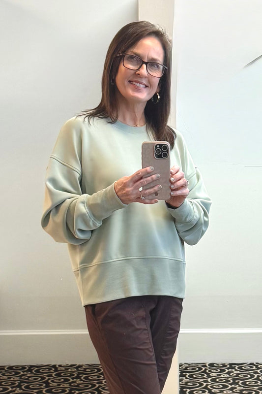Woman wearing a light green sweatshirt and brown pants, holding a phone, standing against a white wall.