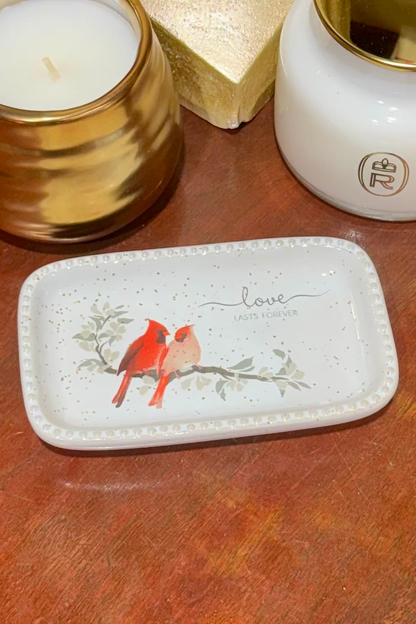 A rectangular white ceramic dish with a beaded rim, featuring a red cardinal and a pink bird perched on a branch with the word 'love' and 'LADY AND MAN' printed on it. The dish is displayed on a wooden surface next to a gold candle and a white candle jar.