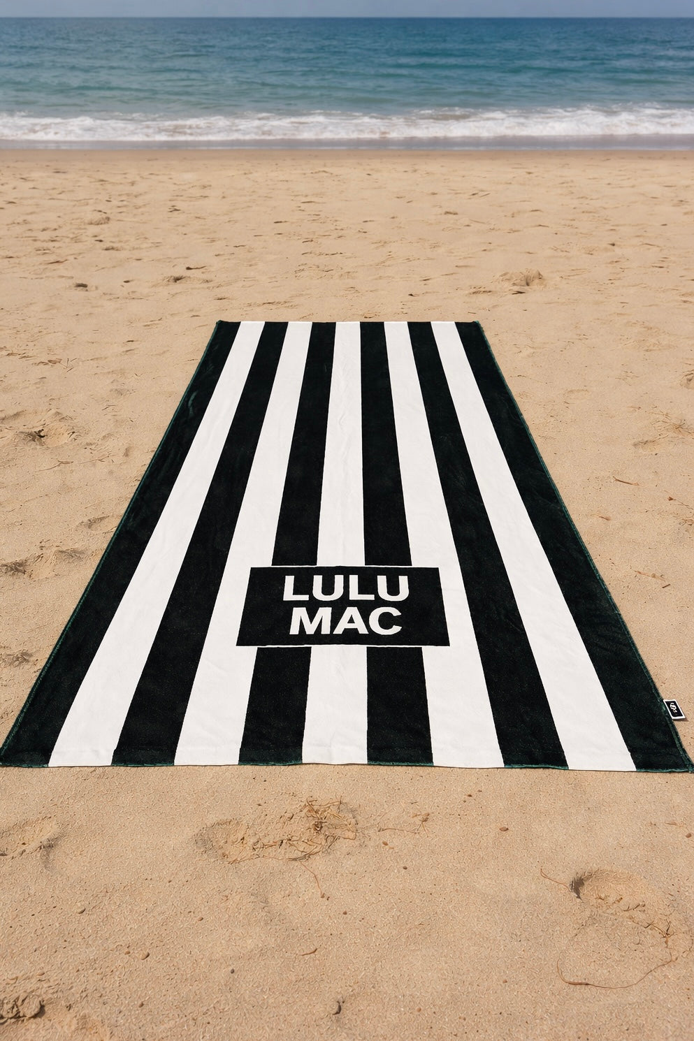 Black and white striped towel with 'Lulu Mac' branding on a sandy beach.