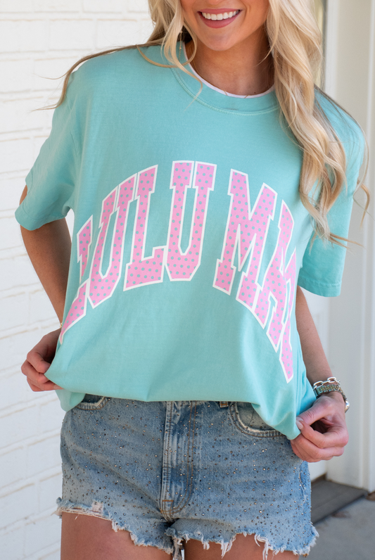 Person wearing a light blue mint lulu Mac t-shirt with pink text and denim shorts against a white brick wall.