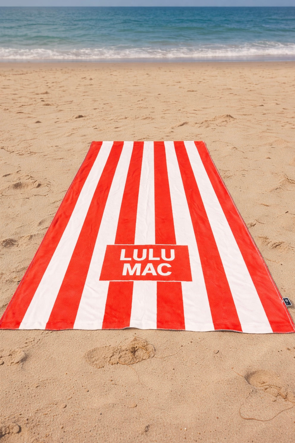 Red and white striped towel with 'Lulu Mac' logo on a sandy beach.