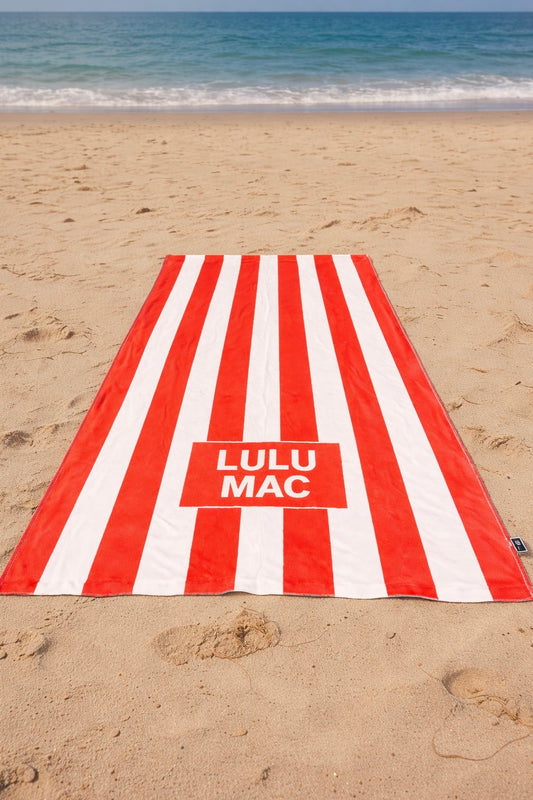 Red and white striped towel with 'Lulu Mac' logo on a sandy beach.
