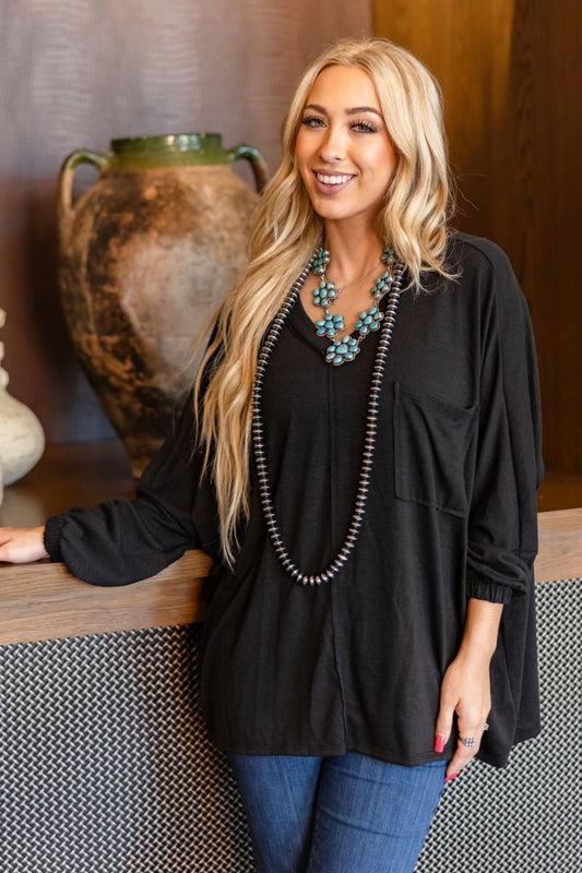 Woman wearing a black top and blue jeans with a necklace, standing indoors.
