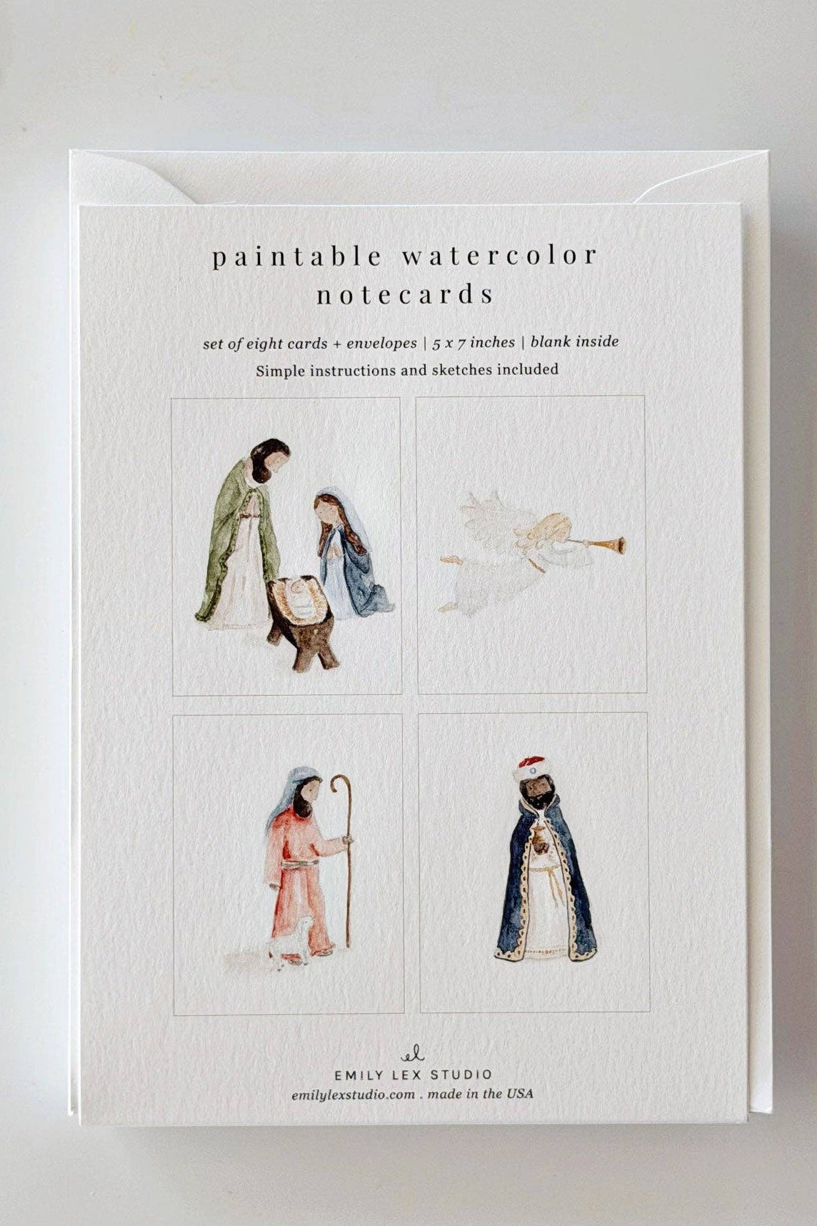 A set of paintable watercolor notecards by Emily Lee Art Studio, displayed on a white surface with a paintbrush and gold star decorations. The notecards are shown with their white envelope, featuring watercolor illustrations of figures and an angel.